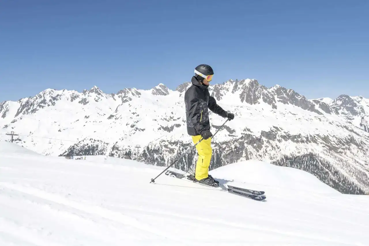 Narciarz w żółtych spodniach i czarnej kurtce z kaskiem i goglami, gotowy do zjazdu. Zastanawiasz się, ile ważą narty zjazdowe?
