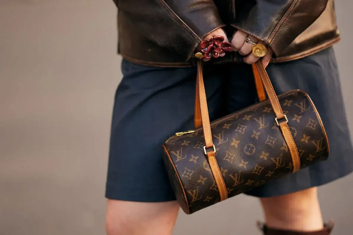 Elegancka, brązowa louis vuitton torebka czarna z monogramem, noszona przez osobę w skórzanej kurtce i granatowej spódnicy.