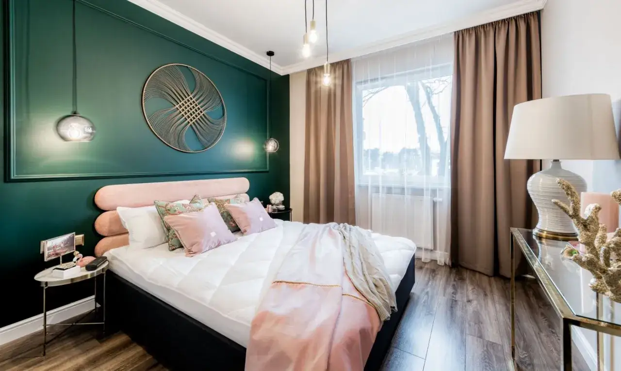 Sypialnia z ł&oacute;żkiem w kolorze pudrowy r&oacute;ż i ścianą w odcieniu butelkowa zieleń. Miękkie tkaniny i złote akcenty tworzą przytulną atmosferę.