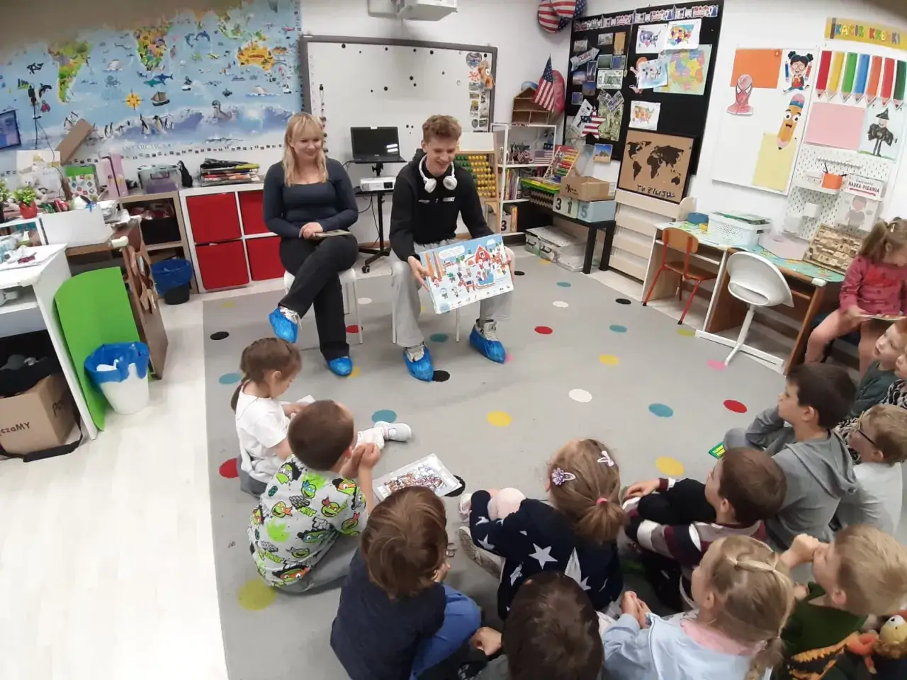 W polskiej klasie przedszkolnej nauczycielka i starszy uczeń czytają dzieciom książkę. Kolorowa sala sprzyja nauce.