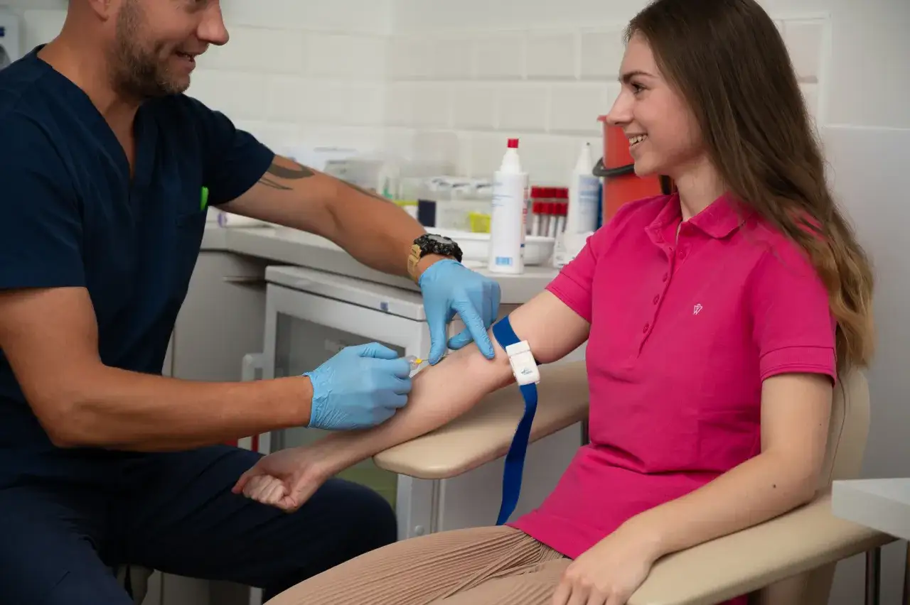 Uśmiechnięta pacjentka podczas pobierania krwi. Pielęgniarz w niebieskich rękawiczkach wykonuje zastrzyk, by pokazać, jak nie zemdleć przy pobieraniu krwi.