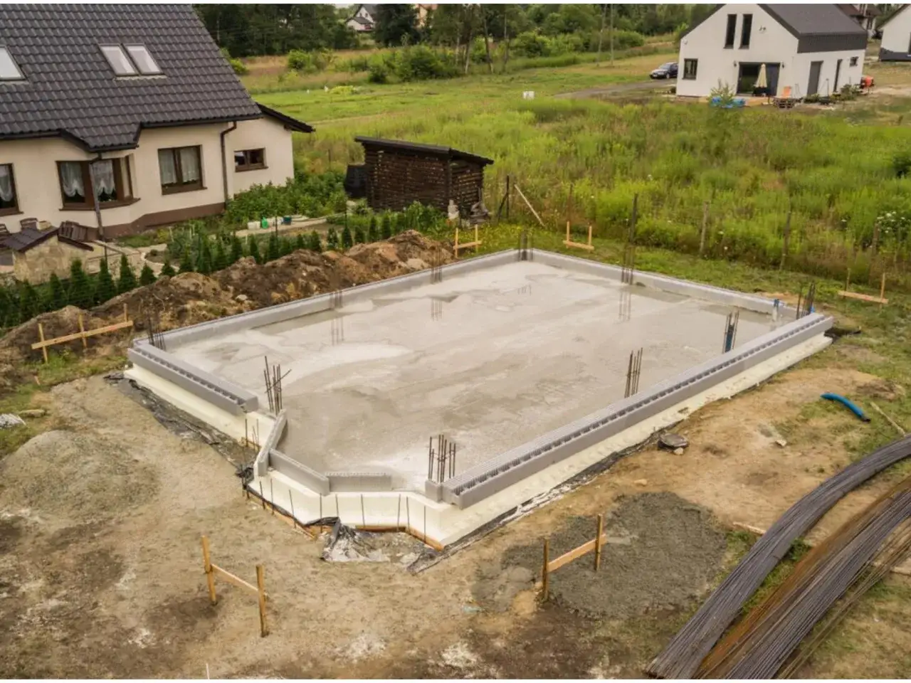 Fundament domu wylewany, gotowy do dalszych prac budowlanych. W pobliżu materiały budowlane i domy. Warto sprawdzić warunki przyłączenia do sieci energetycznej.