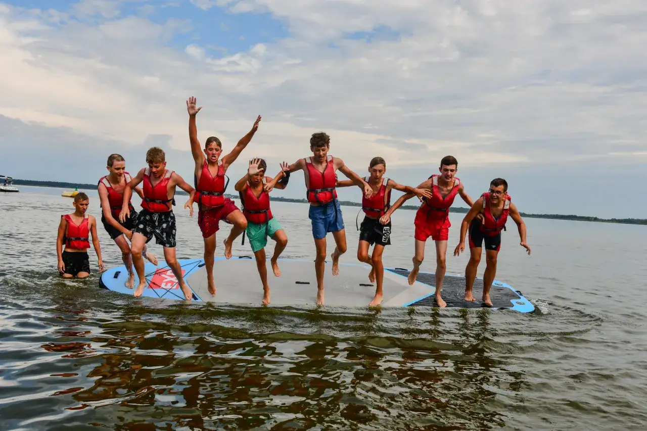 Grupa chłopców w kamizelkach ratunkowych skacze z deski SUP na jeziorze. Dąbki czy warto jechać? Zdecydowanie tak, dla takich chwil!
