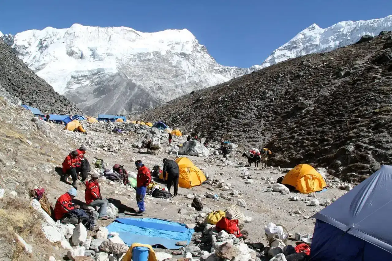 Ob&oacute;z pod ośnieżonym island peak. Ludzie w czerwonych kurtkach odpoczywają wśr&oacute;d namiot&oacute;w, a w tle widać g&oacute;ry.