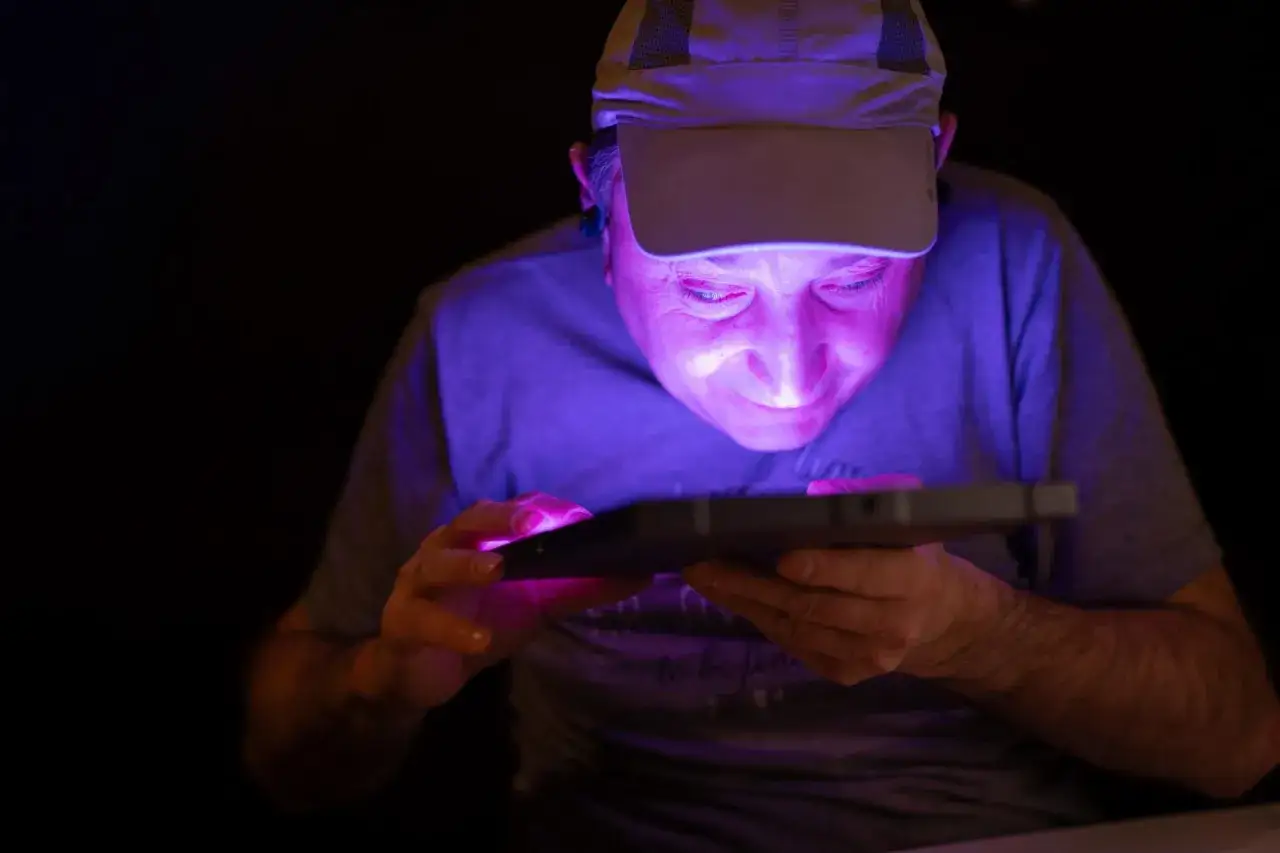 Mężczyzna w czapce z daszkiem pochyla się nad tabletem, jego twarz oświetlona jest fioletowym blaskiem. To jak wystawa w ciemności, gdzie ekran staje się jedynym źródłem światła.