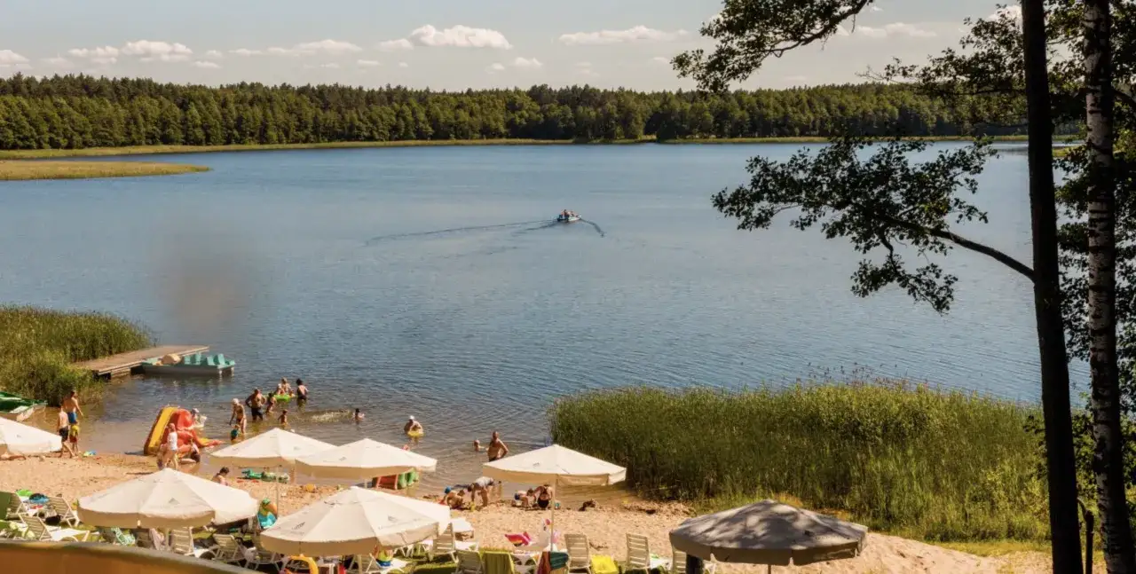 Słoneczny dzień na plaży z parasolami i ludźmi kąpiącymi się w jeziorze. Idealne mazury all inclusive dla całej rodziny.