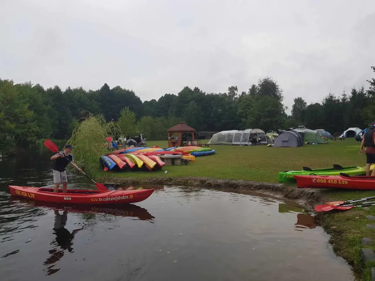 Chłopak w kajaku na rzece Piława, gotowy na spływ. Na brzegu czekają inne kajaki i namioty kempingowe.