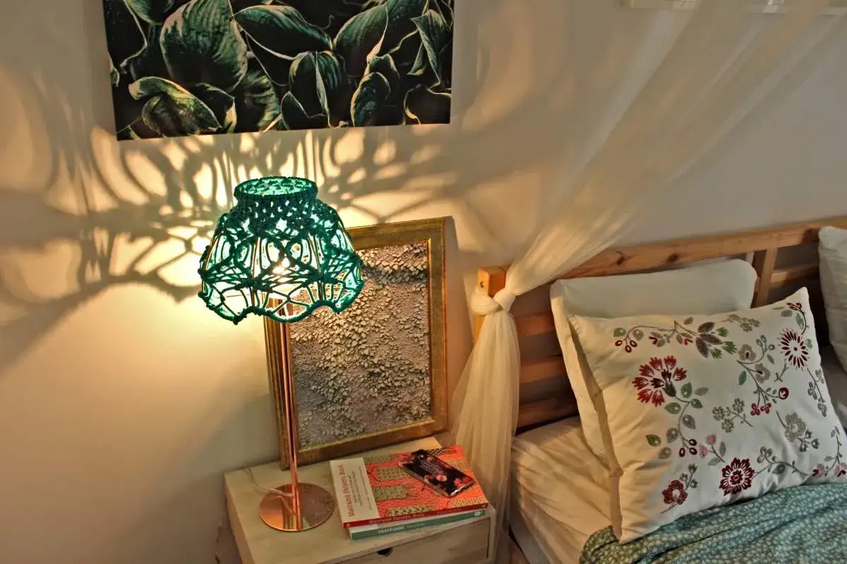 Oświetlona zielona, pleciona lampa rzuca cienie na ścianę. Obok łóżko z kwiecistą poduszką. Jak odnowić stary abażur?