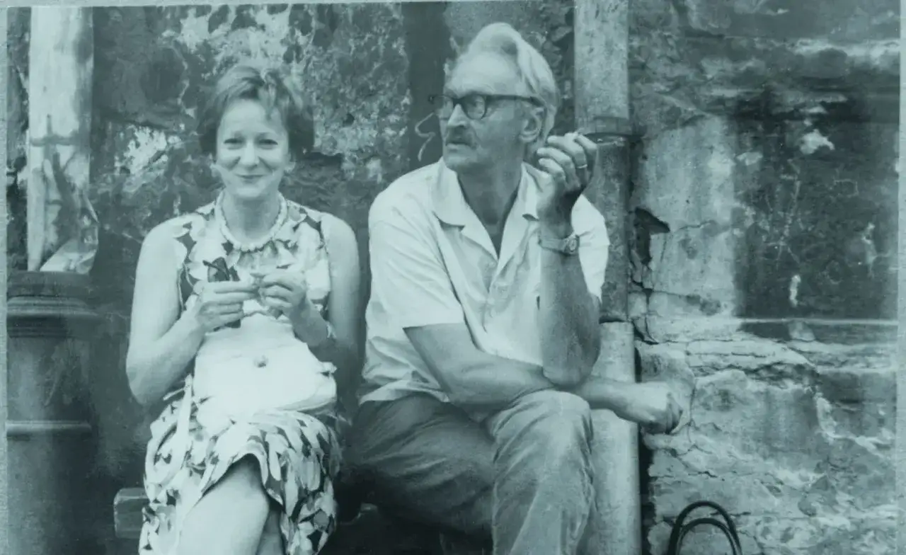 Wisława Szymborska i jej mąż, Adam, siedzą obok siebie. Miłość od pierwszego wejrzenia? Może. Uśmiechnięta poetka trzyma coś w dłoniach, a jej mąż pali papierosa.