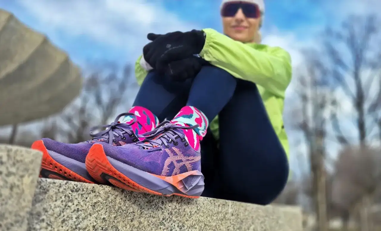 Fioletowe buty do biegania Asics Megablast z pomarańczowymi akcentami na podeszwie. Kobieta w sportowym stroju siedzi na kamiennej ławce.