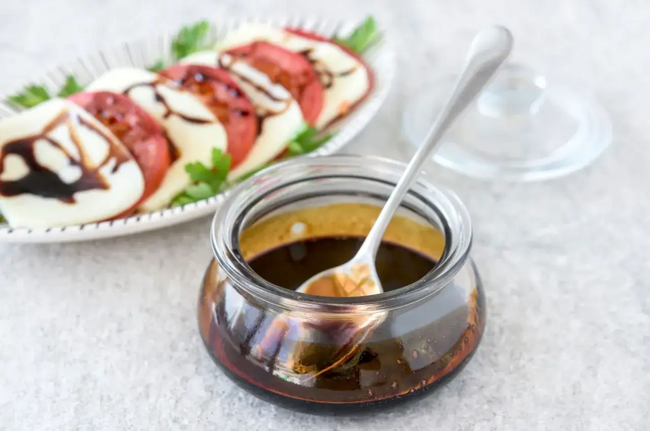 Słoiczek z gęstym, ciemnym sosem balsamicznym i łyżeczką. W tle sałatka caprese z pomidorami, mozzarellą i natką pietruszki, polana tym samym sosem.