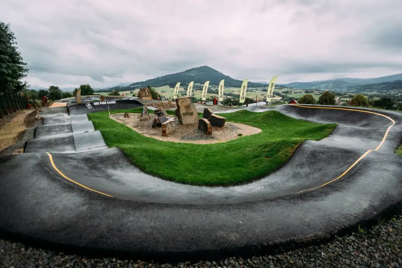 Asfaltowe tory z muldami i bandami w bike park Kasina, otoczone zielenią i kamieniami, z flagami w tle.