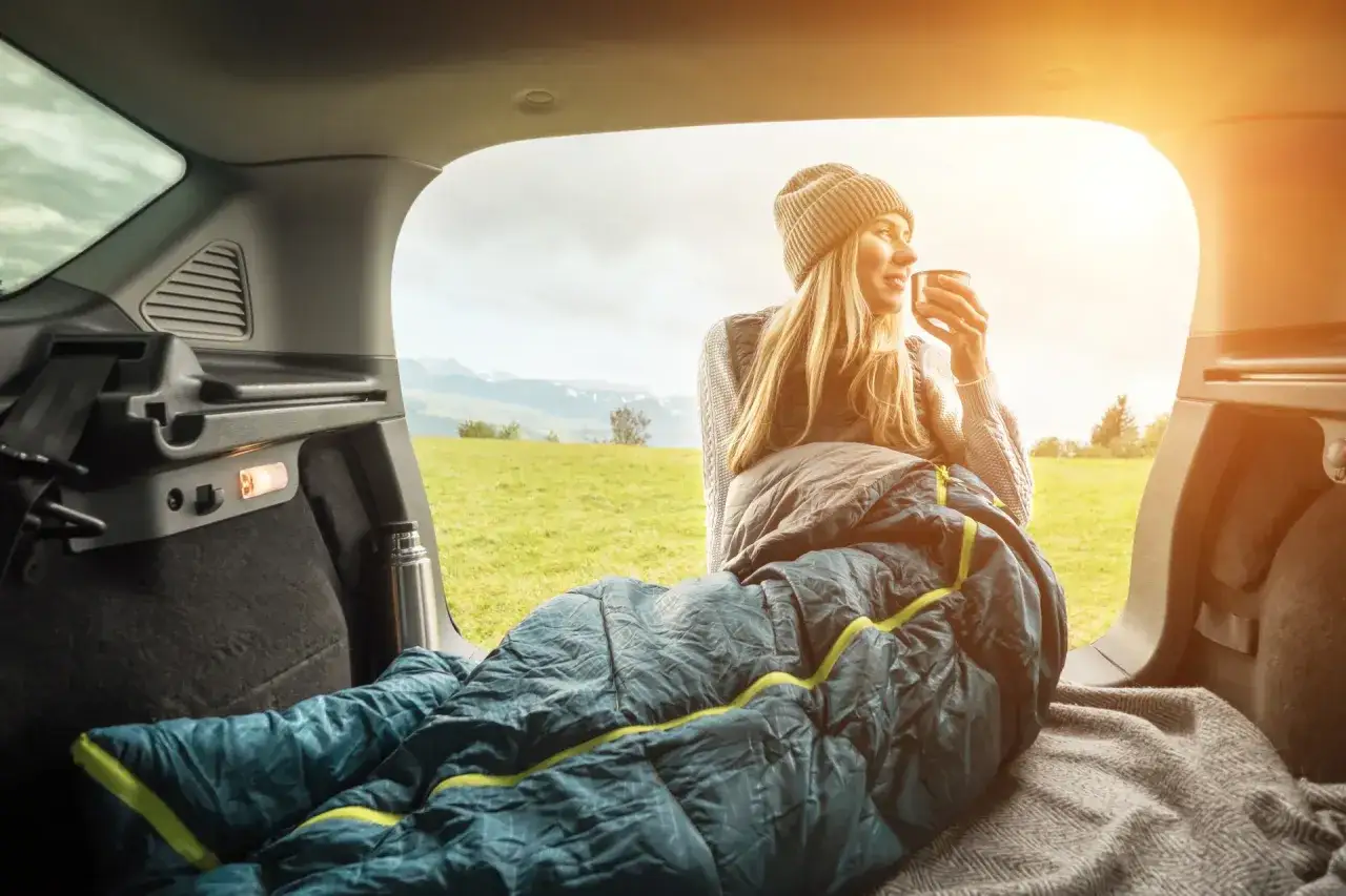 Kobieta pije kawę w śpiworze w bagażniku samochodu, ciesząc się widokiem g&oacute;r. To idealne rozwiązanie na spanie w samochodzie w Europie.