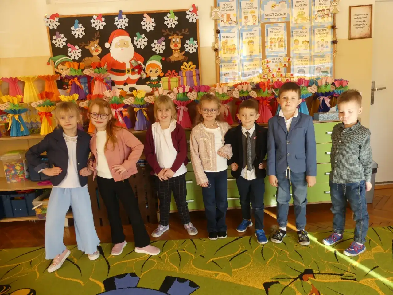 Dzieci w przedszkolu, ubrane odświętnie, pozują na tle świątecznych dekoracji. Czy przedszkole ma ferie? Nie, widać radość i przygotowania do świąt.