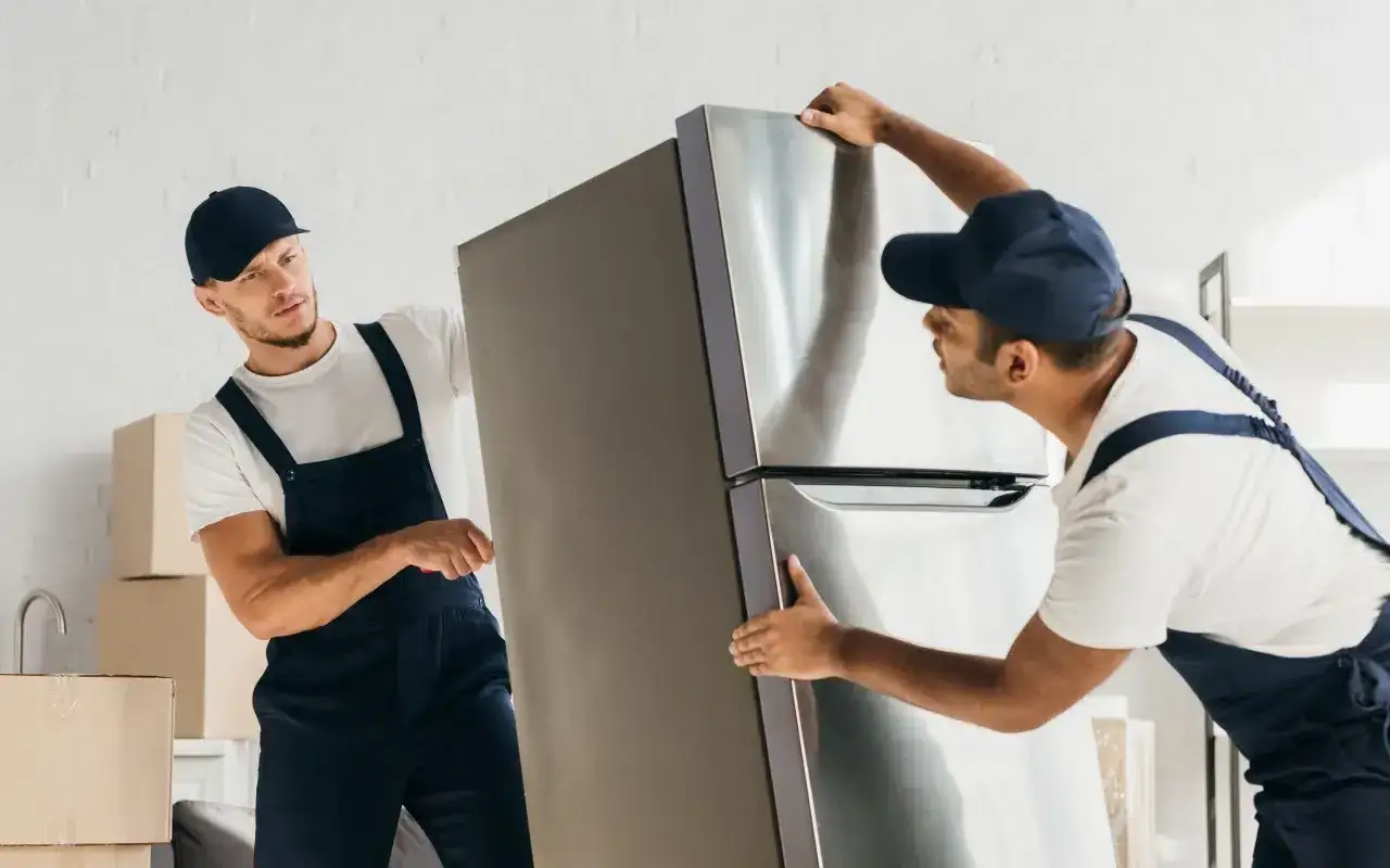 Dw&oacute;ch mężczyzn w niebieskich ogrodniczkach ostrożnie przenosi lod&oacute;wkę, pokazując, jak przewozić ten sprzęt AGD.