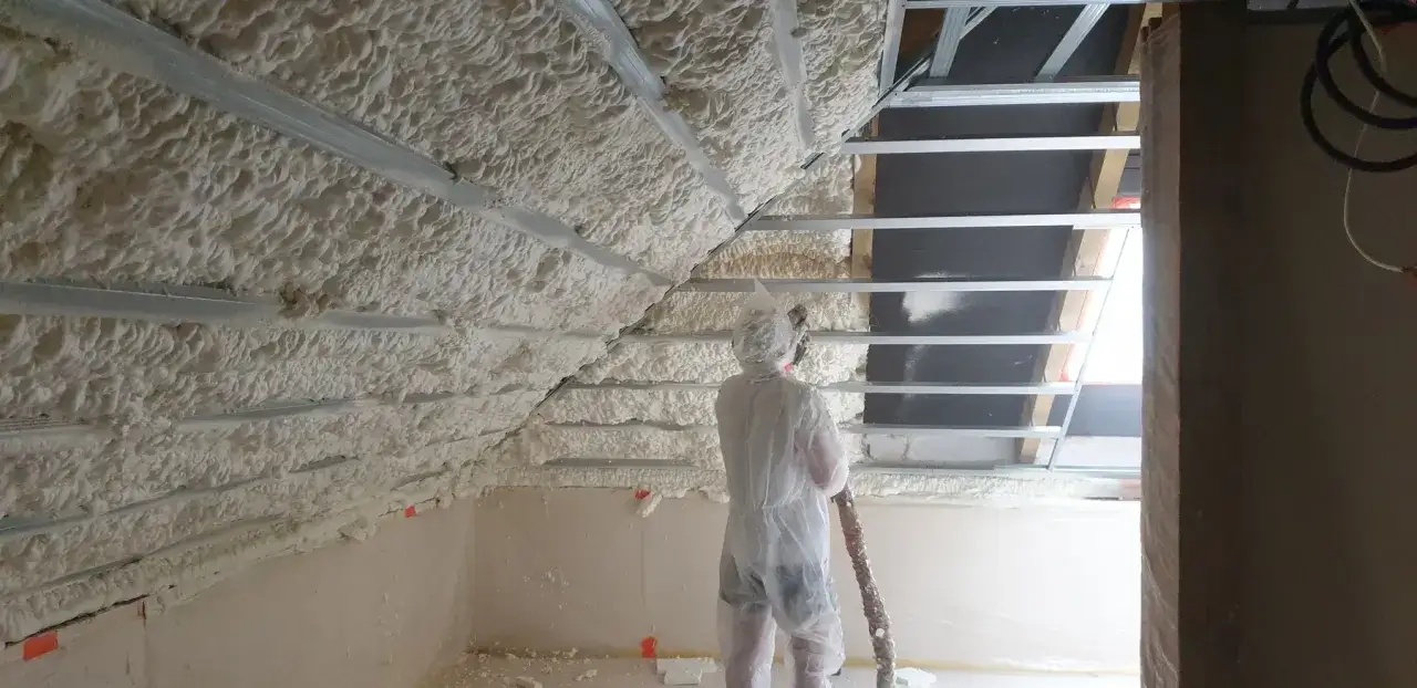 Pracownik w kombinezonie wykonuje ocieplanie pianką PUR poddasza. Pianka pokrywa konstrukcję dachu, tworząc jednolitą warstwę izolacyjną.