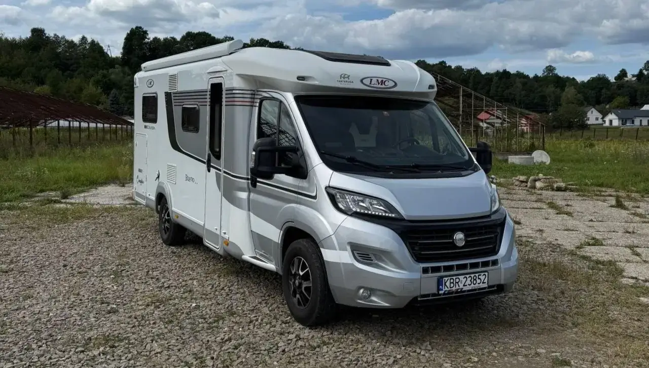 Nowy kamper 4 osobowy LMC, gotowy na rodzinne przygody. Biało-srebrny, z nowoczesnym designem, zaparkowany na żwirowym podjeździe.