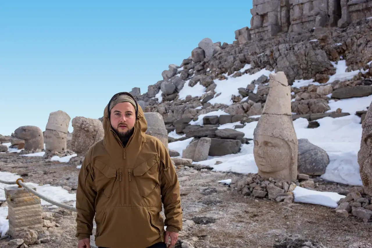 Mężczyzna w brązowej kurtce na tle starożytnych posąg&oacute;w na g&oacute;rze Nemrut. Śnieg i kamienie tworzą surowy krajobraz.