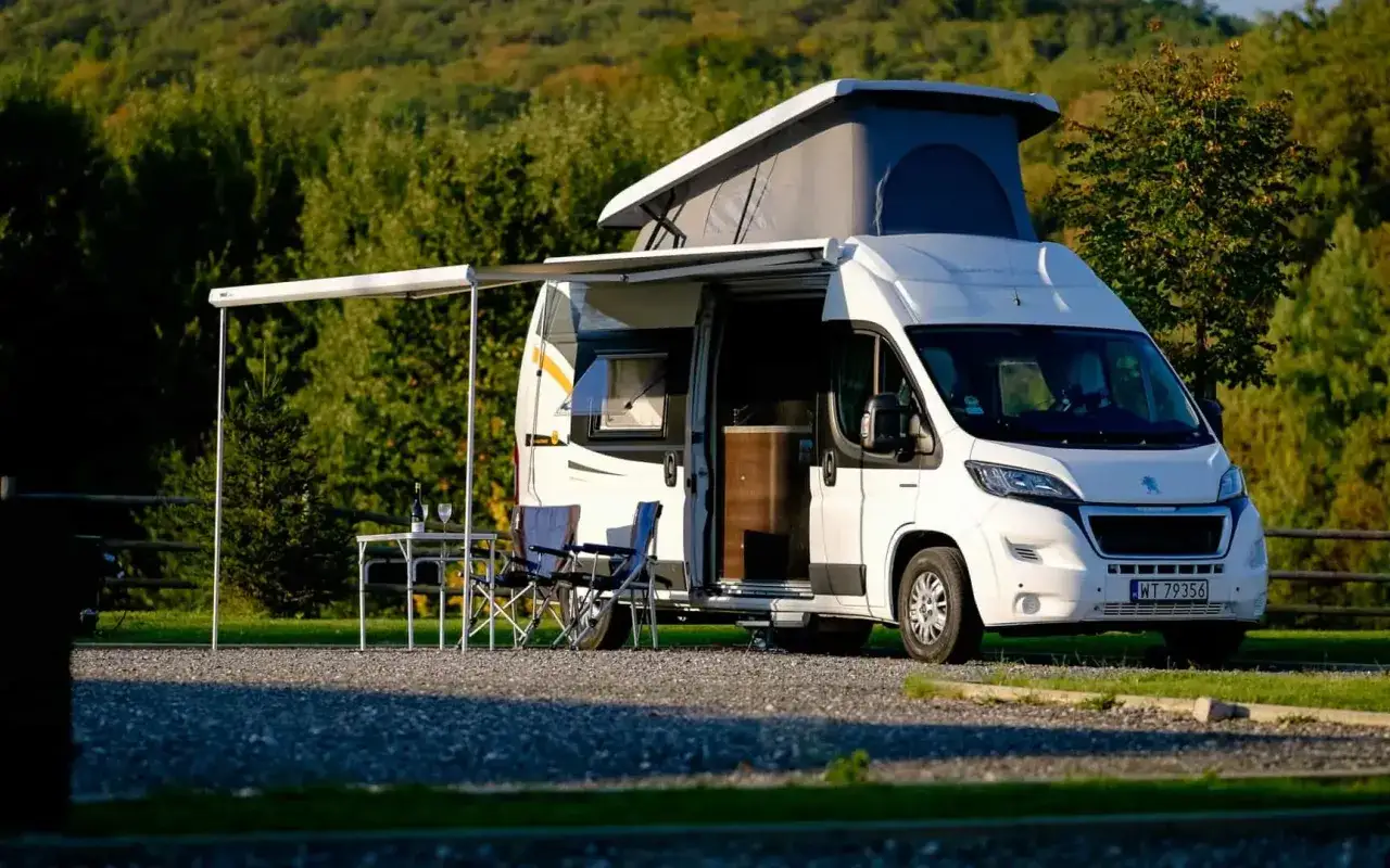 Biały kamper z podnoszonym dachem, rozłożonym markizą i stolikiem z krzesłami, gotowy na wakacje.