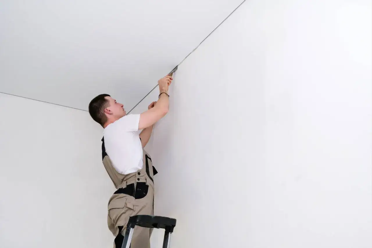 Wykończenie narożnika sufitu. Fachowiec na drabinie wykonuje precyzyjne nakładanie akrylu przy suficie, zapewniając idealnie gładką linię.