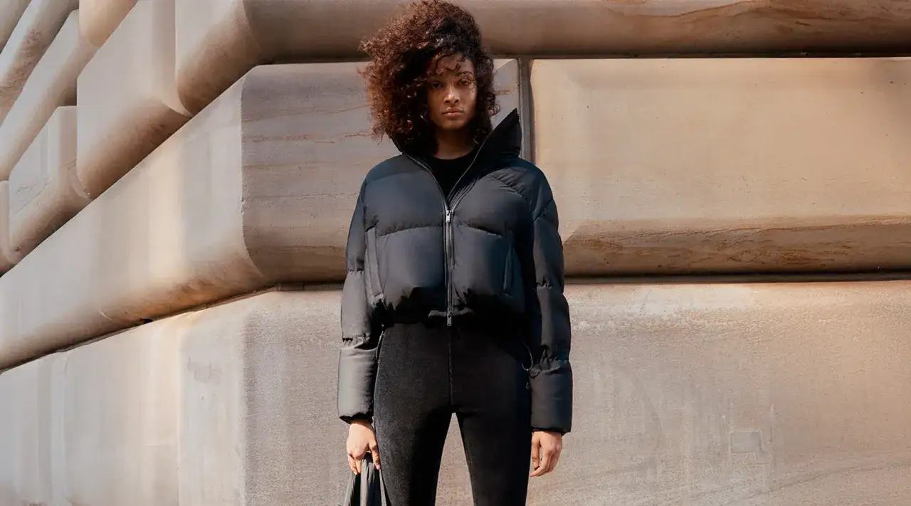 Kobieta w czarnej, puchowej kurtce prezentuje modne kurtki na jesień. Stylizacja uzupełniona czarnymi spodniami i torebką.