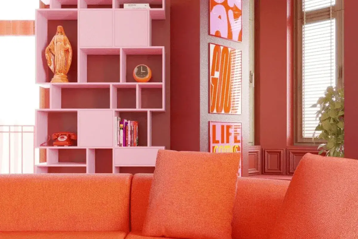 Pomarańczowa sofa i r&oacute;żowe regały tworzą radosny dopamine decor. Na p&oacute;łce figurka, zegar i książki. Obok plakaty z napisami.