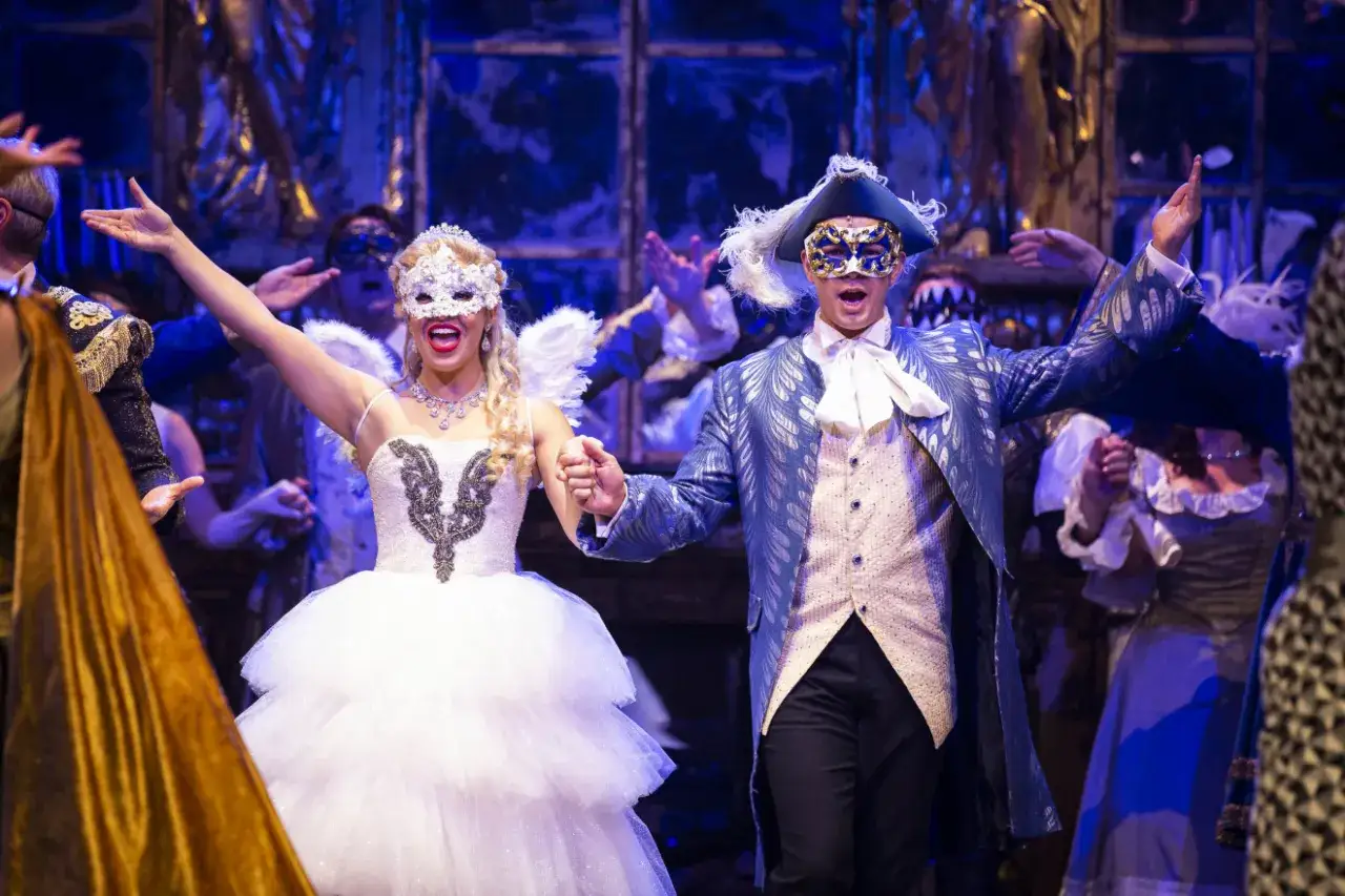 Upiór w operze obsada: para w maskach i strojach balowych, ona w białej sukni z anielskimi skrzydłami, on w niebieskim fraku.