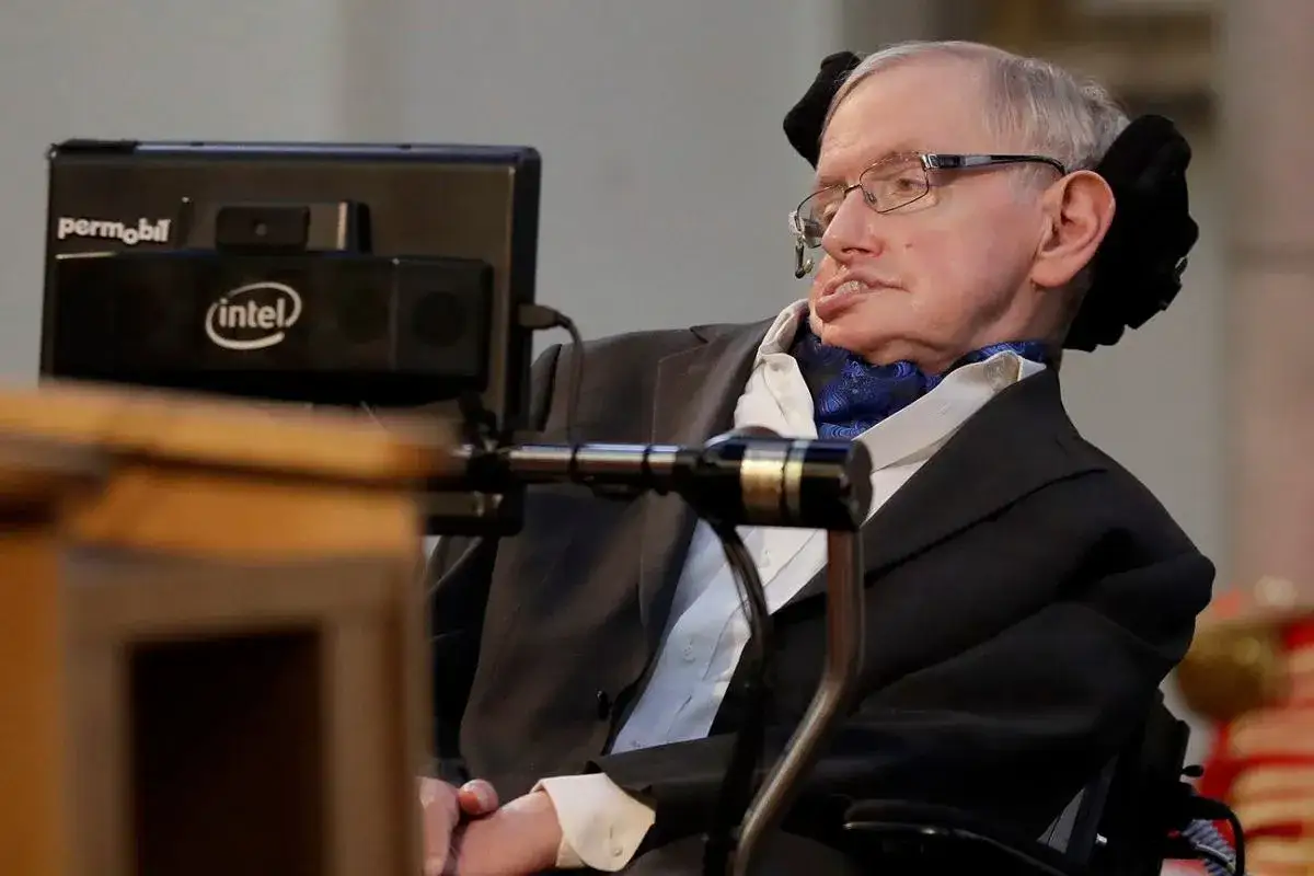 Stephen Hawking, wybitny naukowiec na wózku, w garniturze i z niebieską z aksamitną apaszką, przed komputerem z logo Intel.