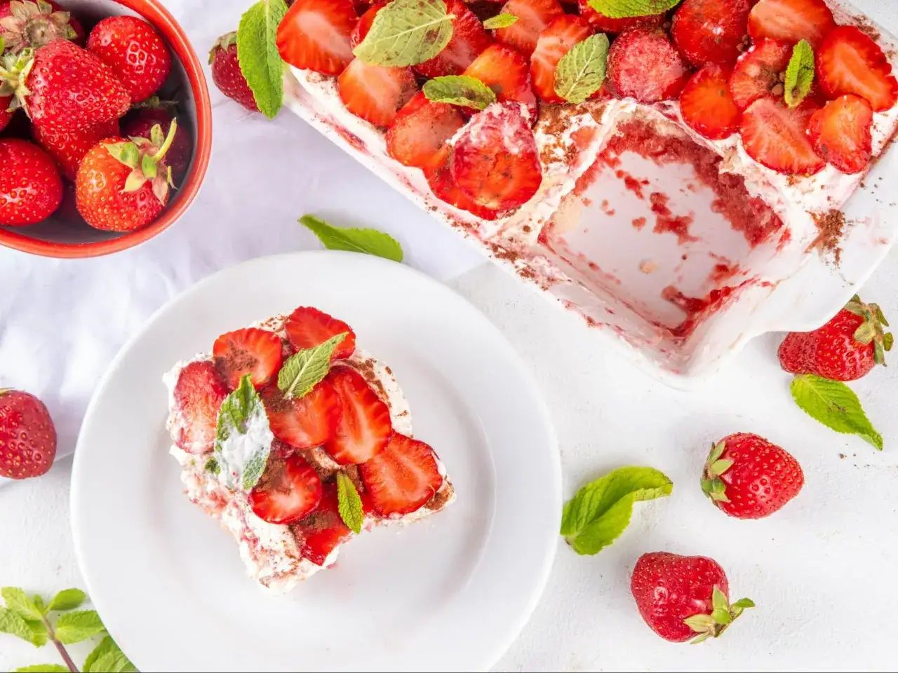 Tiramisu klasyczne kontra tiramisu truskawkowe. Porcja deserowa z truskawkami i miętą na talerzu, obok miseczka owoców i reszta ciasta w naczyniu.
