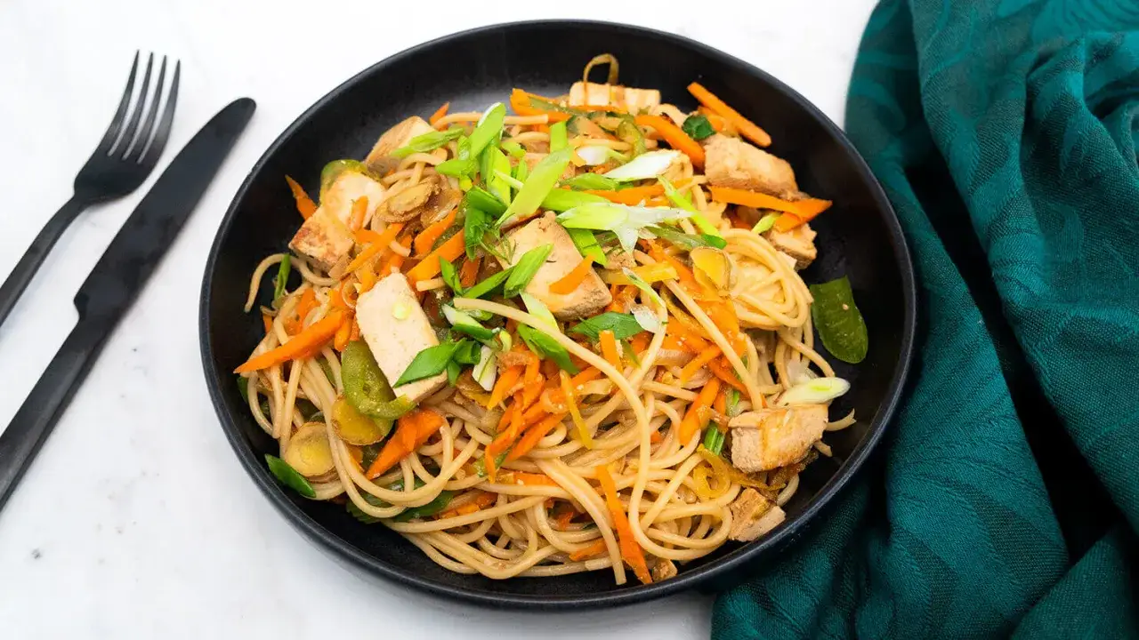 Pyszne makaron z tofu, marchewką i zieloną cebulką w czarnej misce, obok czarnego sztućca i zielonej serwetki.