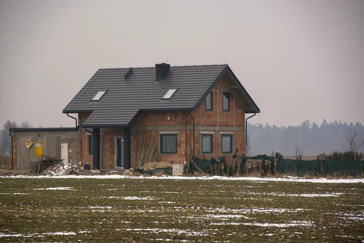 Dom w budowie, z cegły, z szarym dachem, na tle lasu. Widać plac budowy, materiały budowlane i tablicę informacyjną. To baza pozwoleń na budowę.