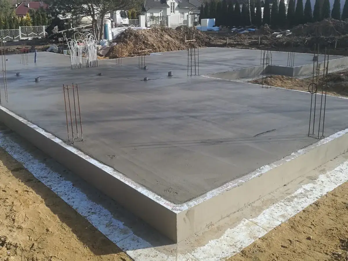 Świeżo wylana płyta fundamentowa z zbrojeniem. Wokół widoczne są elementy, które posłużą do tego, czym obsypać fundamenty, by je zabezpieczyć.