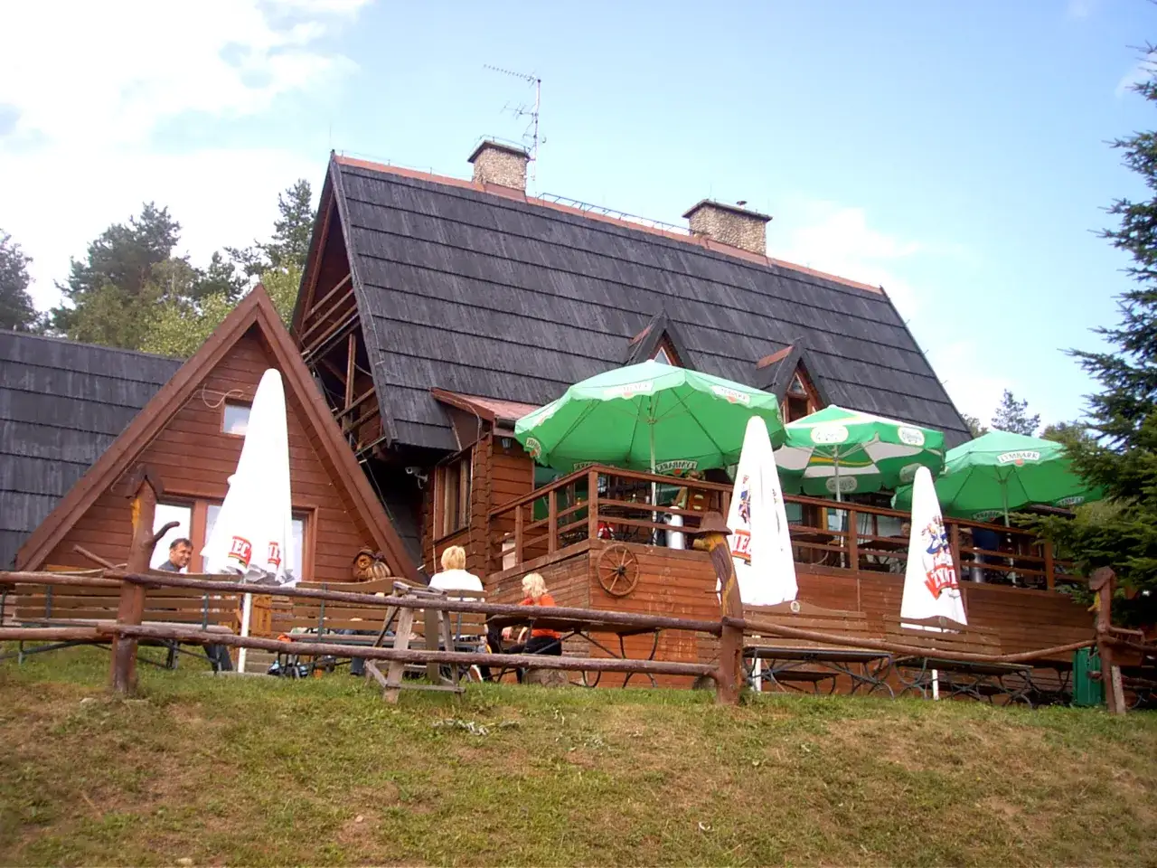 Drewniana bacówka na Jamnej z zielonymi parasolami i ławkami, gdzie odpoczywają ludzie.