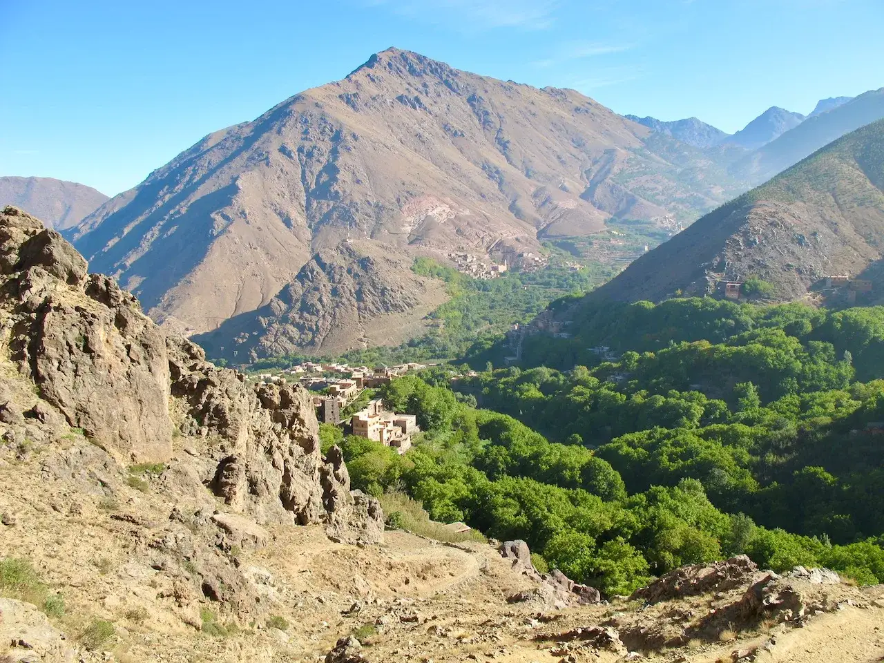 Widok na dolinę z wioskami u podn&oacute;ża masywu Jabal Toubkal. Zielone drzewa kontrastują z surowymi, skalistymi zboczami g&oacute;r.