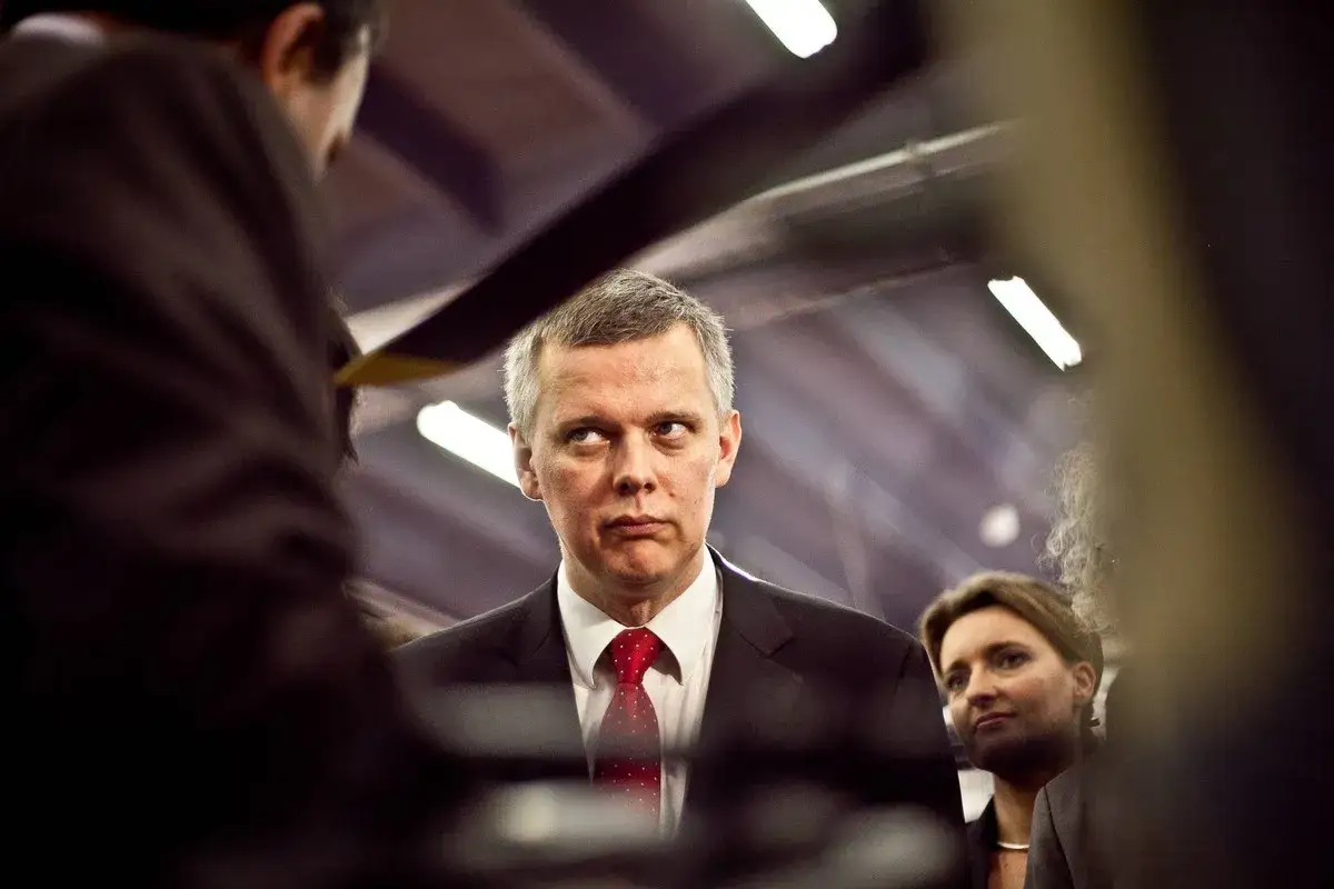 Tomasz Siemoniak, zamyślony, w garniturze i czerwonym krawacie, rozważa swoje pochodzenie. Obok kobieta.