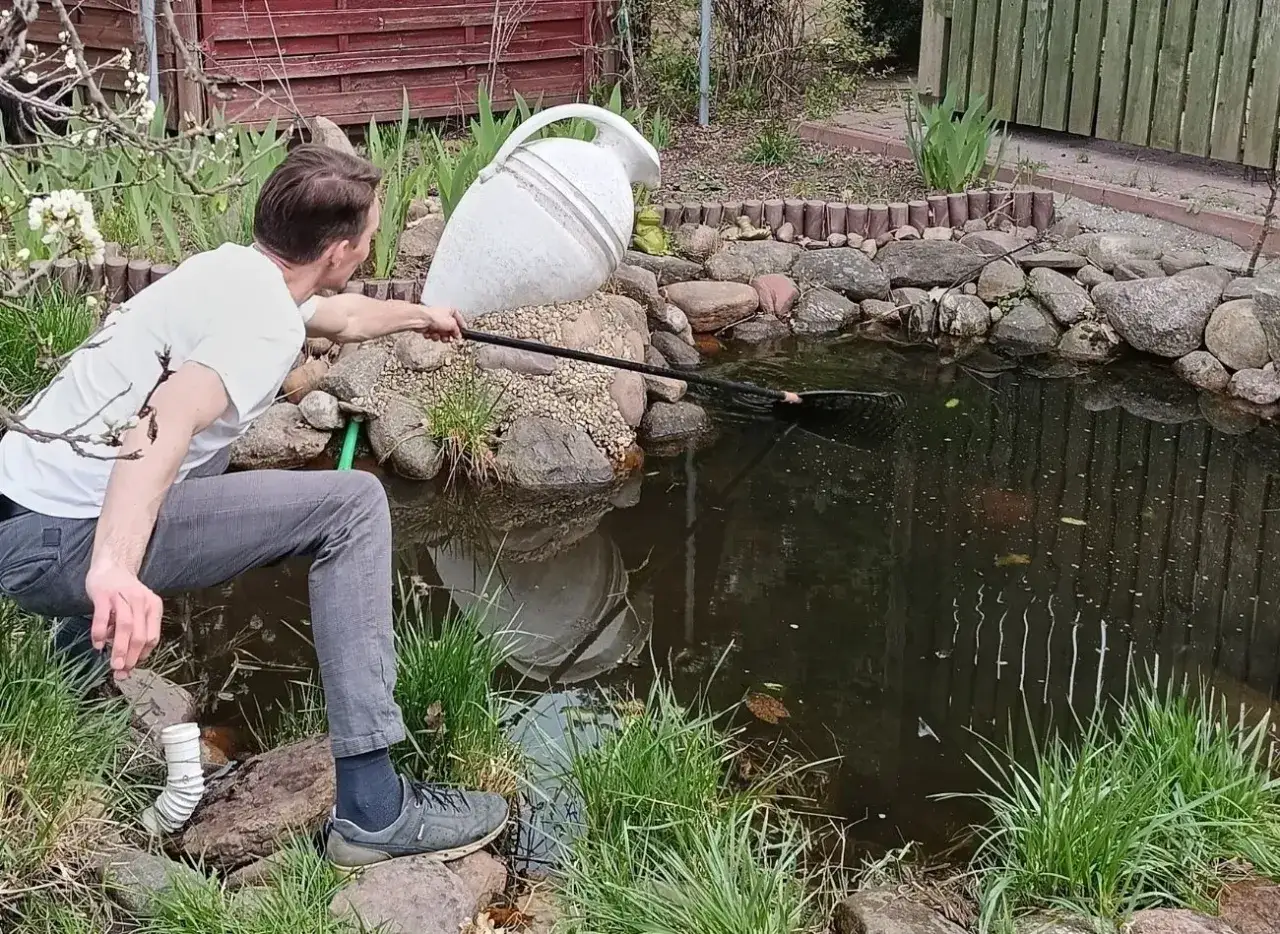 Mężczyzna z podbierakiem wykonuje czyszczenie oczka wodnego. W tle duża, biała amfora.