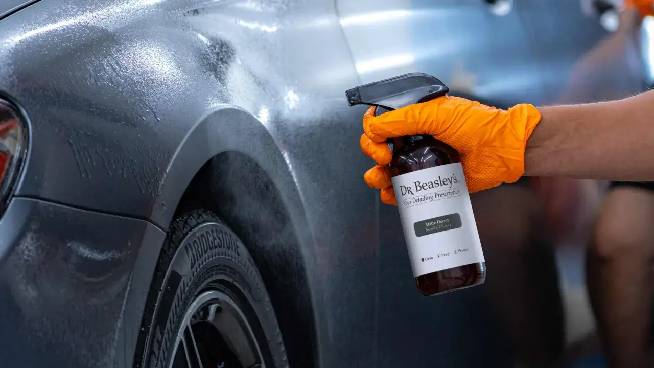 Ręka w pomarańczowej rękawiczce rozpyla preparat Dr. Beasley's Matte Decon na lakierze samochodu, rozpoczynając proces dekontaminacji.