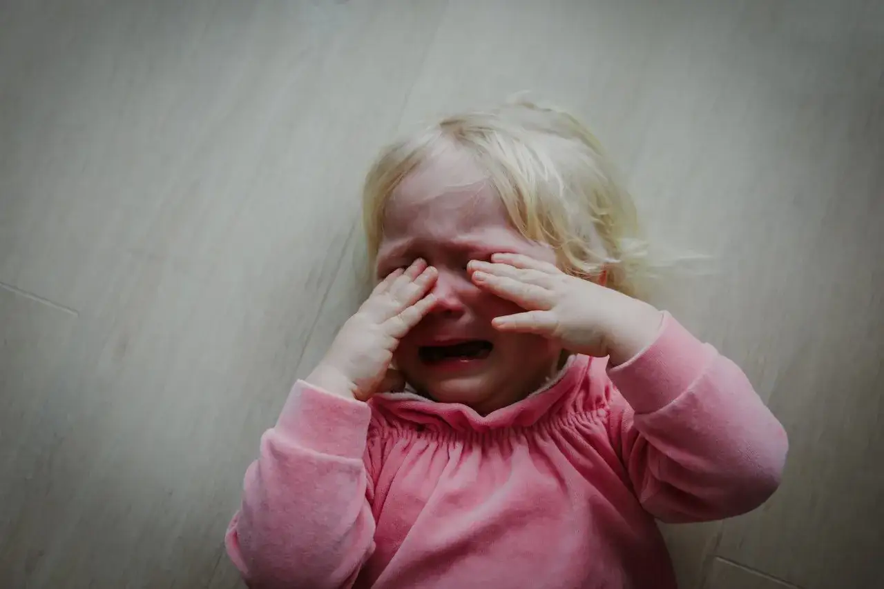 Małe dziecko płacze, zasłaniając oczy rękami. Widać, że dziecko bije się po głowie, wyrażając swoje niezadowolenie.