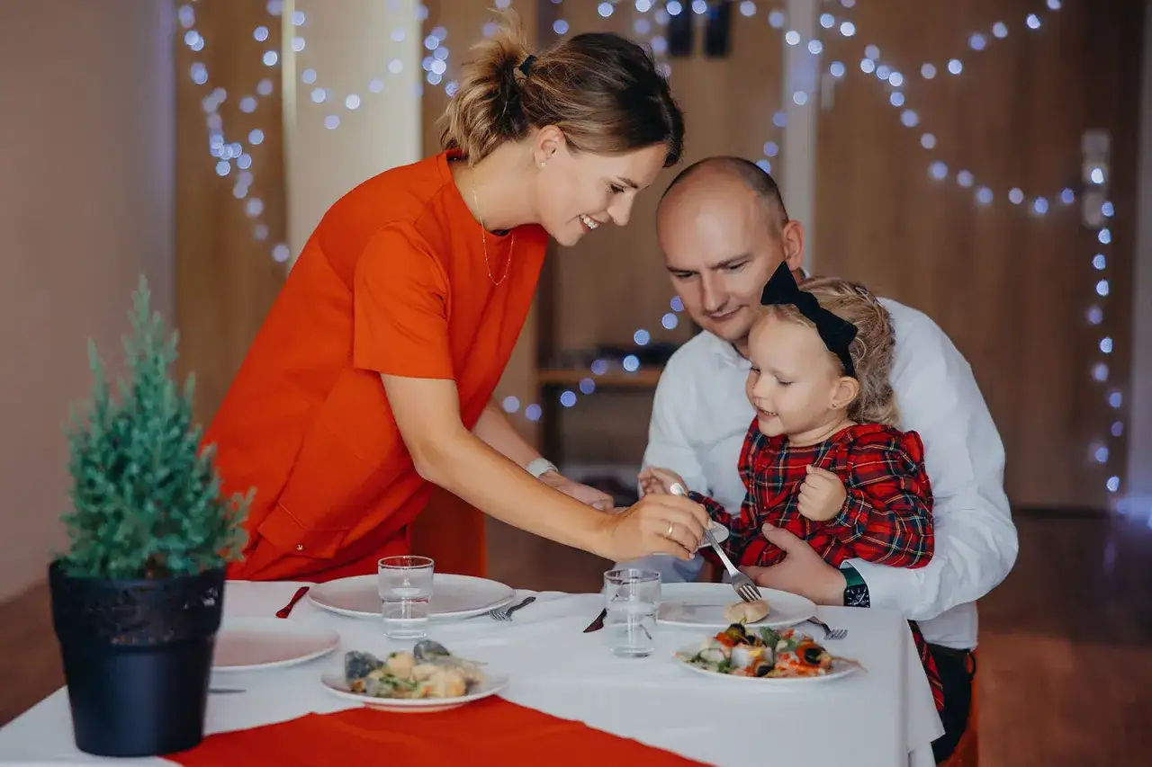 Rodzina celebruje święta w hotelu. Mama karmi córeczkę, tata patrzy z uśmiechem. W tle świąteczne lampki.