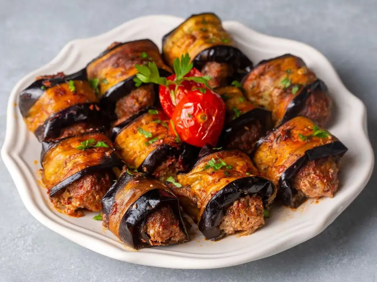Roladki z bakłażana z mięsem mielonym, polane sosem i posypane natką pietruszki. Obok pieczony pomidorek.