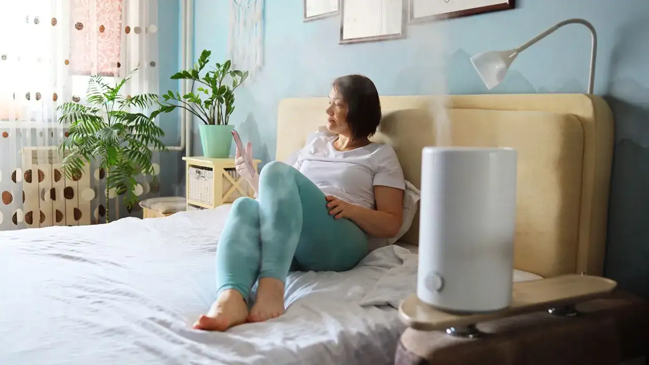 Kobieta na łóżku korzysta z telefonu, obok działa nawilżacz ewaporacyjny, co to znaczy? Lepsze powietrze w pokoju.