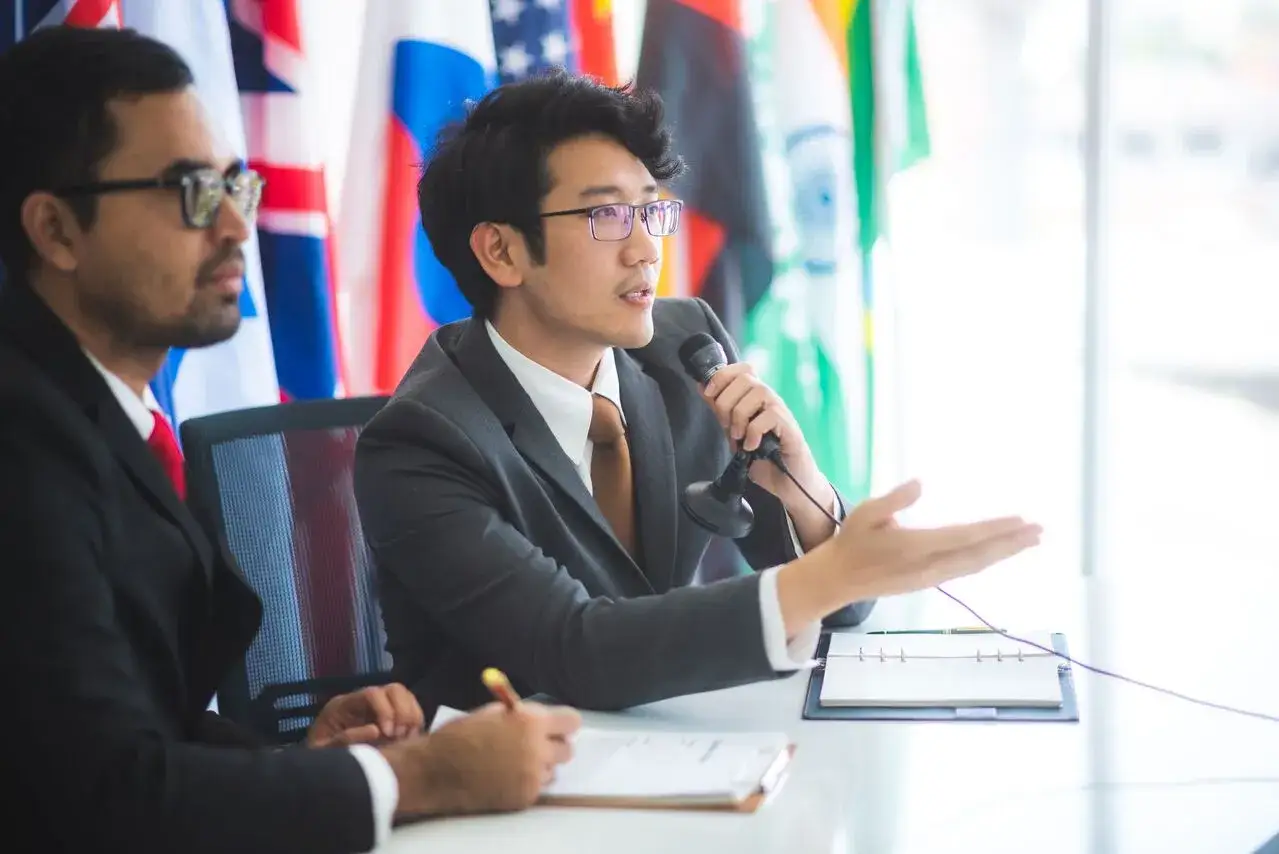 Mężczyzna z mikrofonem, mówiący o tłumaczeniu konsekutywnym a symultanicznym, na tle flag.