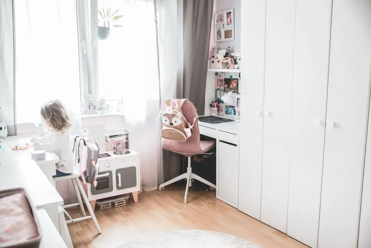 Mały pokój dziecięcy w bloku. Dziewczynka bawi się przy biurku, obok stoi różowa kuchnia i szafa.