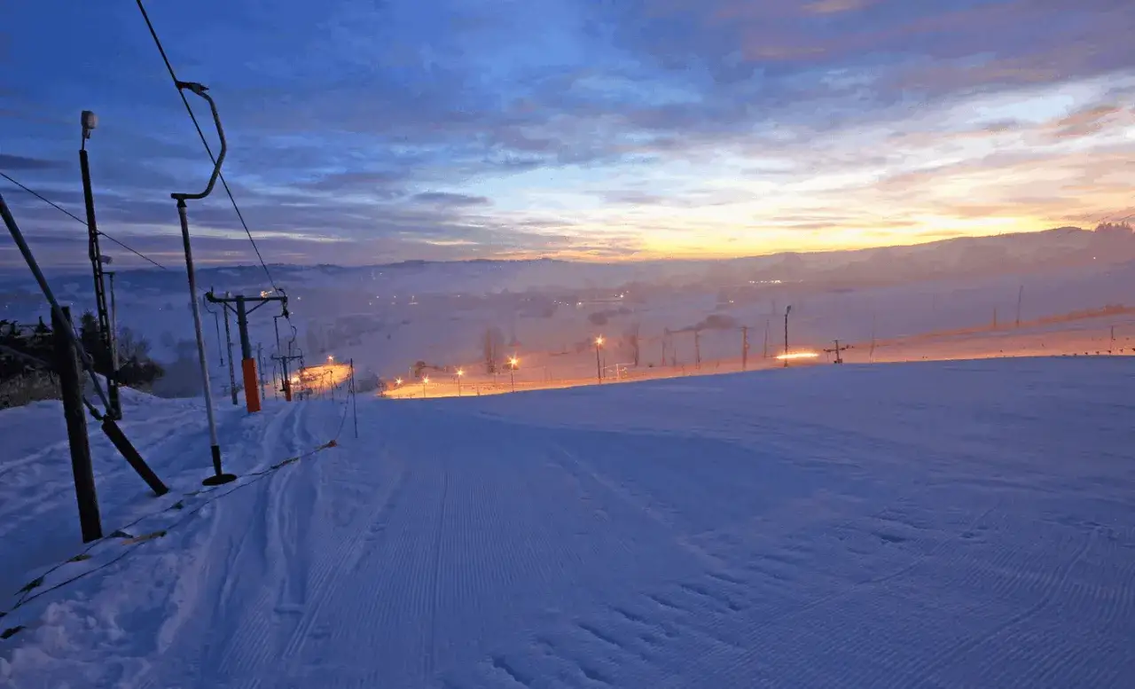 Ośnieżony stok narciarski na Olczańskim Wierchu o wschodzie słońca, z wyciągiem krzesełkowym i latarniami.