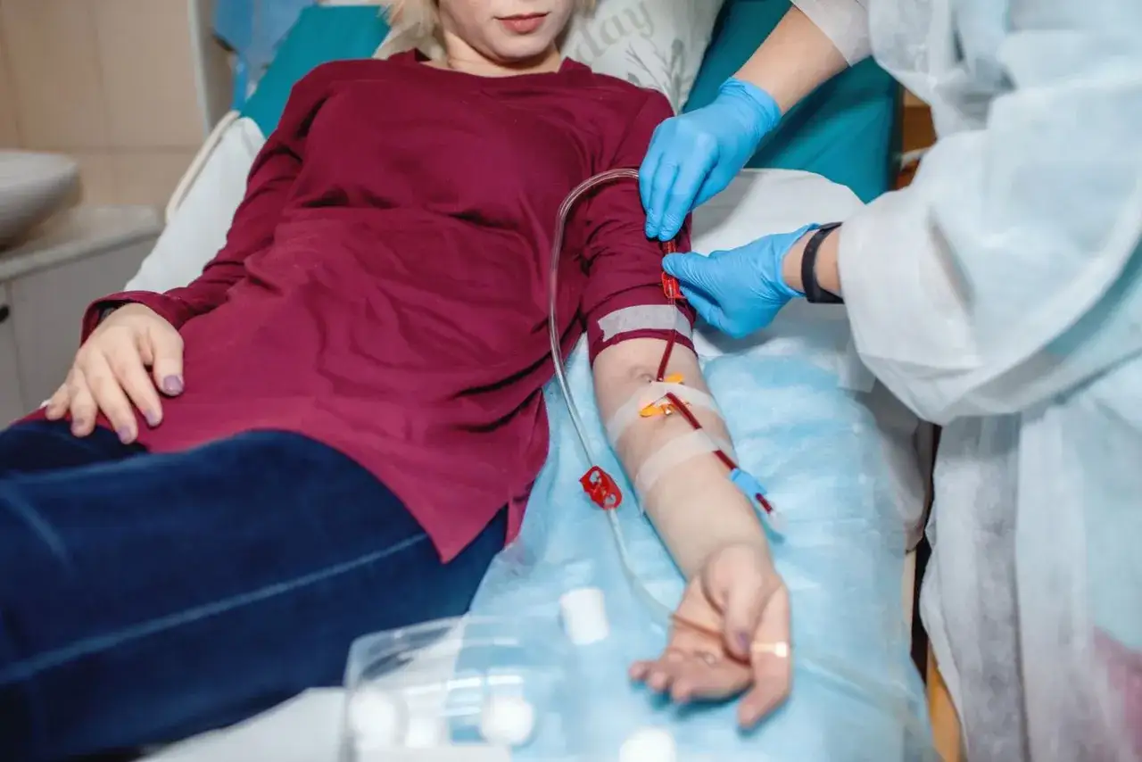 Pacjentka leży na ł&oacute;żku, pielęgniarka w rękawiczkach podłącza przetokę do dializ. Krew płynie przez przezroczyste rurki.