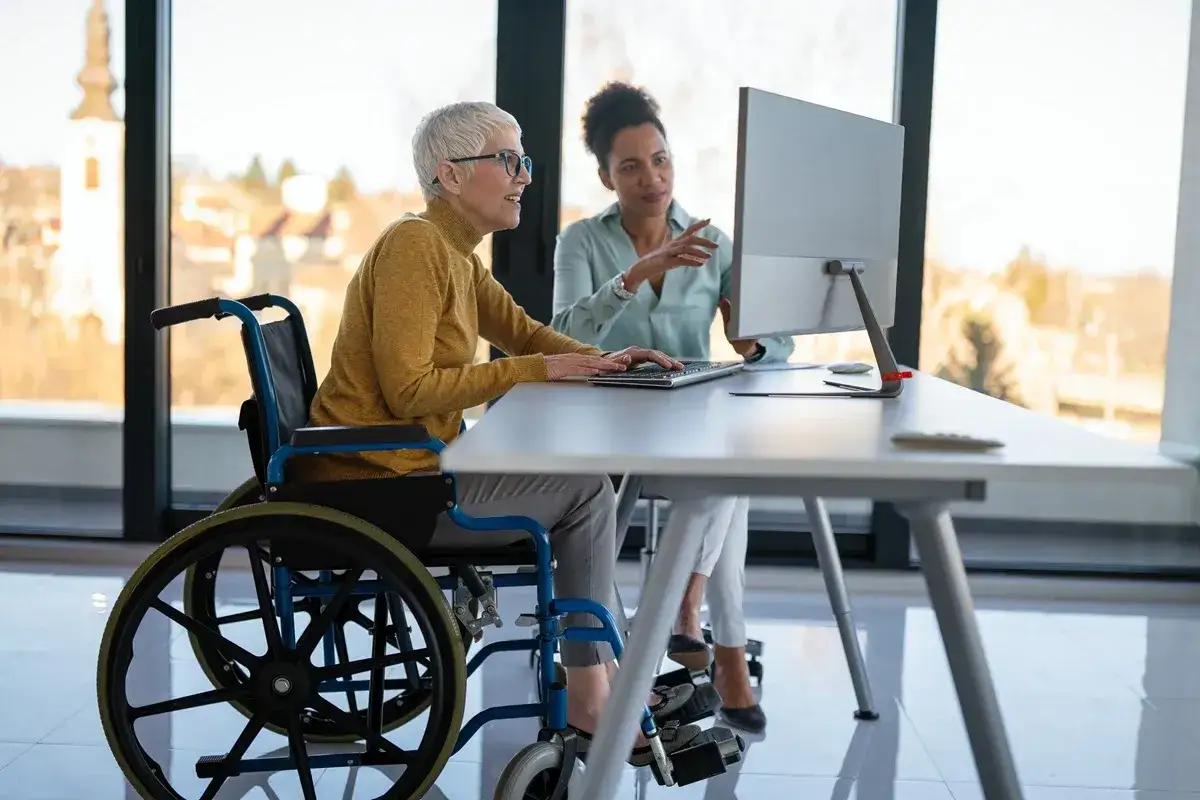 Kobieta na wózku inwalidzkim pracuje przy komputerze z koleżanką. Dyskutują, czy dodatkowy urlop dla osoby niepełnosprawnej przechodzi na następny rok.