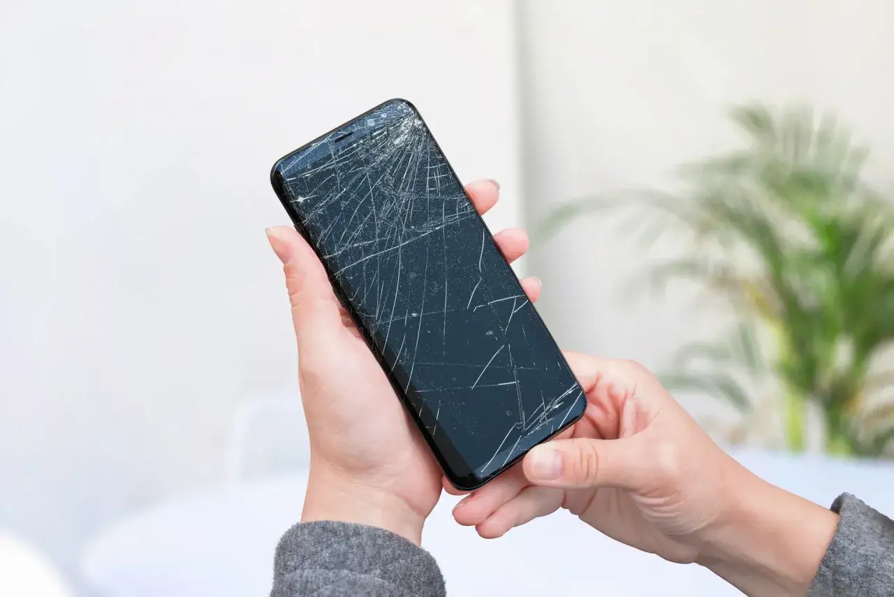 Dłonie trzymają telefon z mocno pękniętą szybką. Czy można używać telefonu z pękniętą szybką? To pytanie zadaje sobie wiele os&oacute;b.