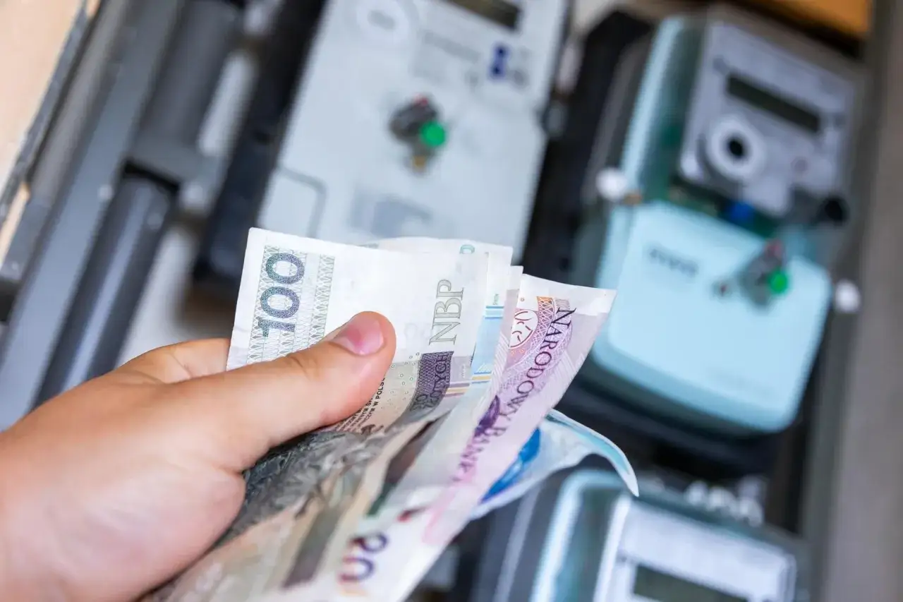 Dłoń trzyma polskie banknoty na tle licznik&oacute;w energii. Zastanawiasz się nad zniżki na prąd dla emeryt&oacute;w wniosek?