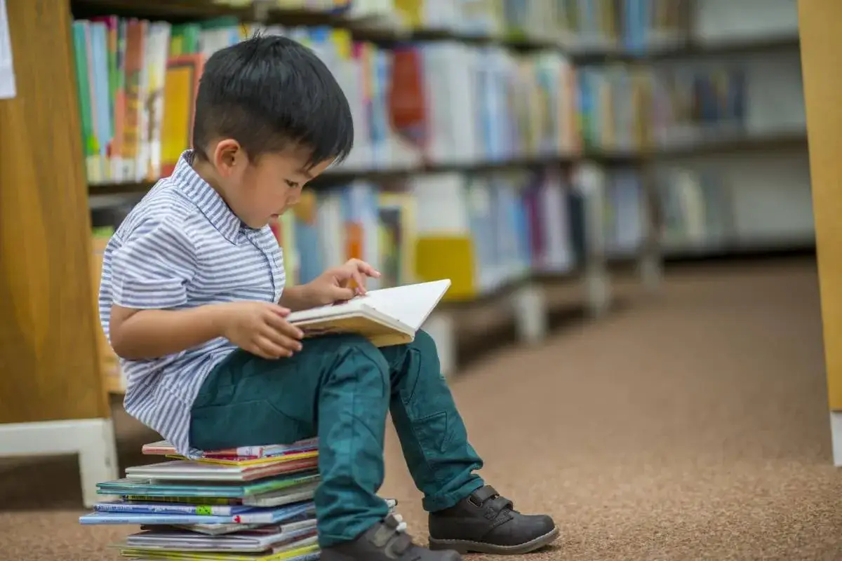 Mały chłopiec siedzi na stosie książek w bibliotece, pochłonięty czytaniem.