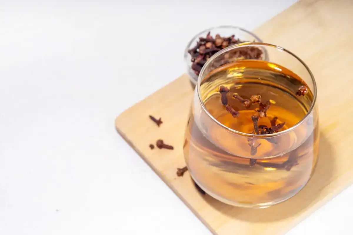 Szklanka z naparem, w której pływają całe goździki. Obok miseczka z suszonymi goździkami.